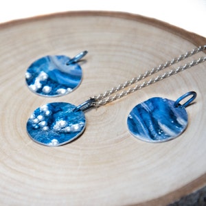 May include: A set of three silver pendant necklaces with blue and white abstract designs. The pendants are round and have a textured surface. The necklaces are on a wooden surface.
