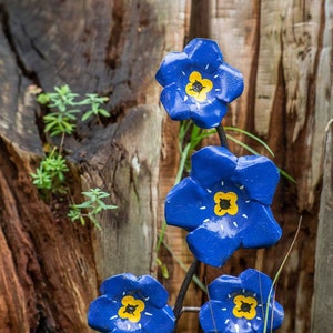 K&ouml;nnte beinhalten: Eine Metallskulptur einer Blume mit blauen Bl&uuml;tenbl&auml;ttern und gelben Zentren. Die Skulptur ist so gestaltet, dass sie wie eine Vergissmeinnicht-Blume aussieht.
