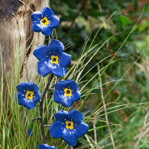 K&ouml;nnte beinhalten: Eine Metallskulptur eines Blumenst&auml;ngels mit blauen Bl&uuml;ten und gelben Zentren. Die Bl&uuml;ten sind in einer Traube am Stiel angeordnet. Die Skulptur ist vor einem Hintergrund aus gr&uuml;nem Gras platziert.