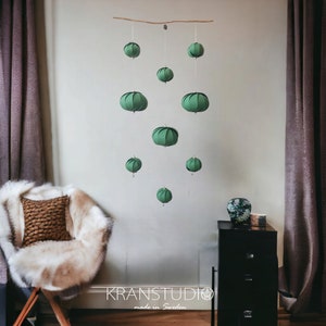 Peut inclure: Un mobile en papier vert avec 9 ornements ronds suspendus. Le mobile est suspendu à une branche et pend devant un mur blanc. Le texte "KRANSTUDIO made in Sweden" est visible en bas de l'image.