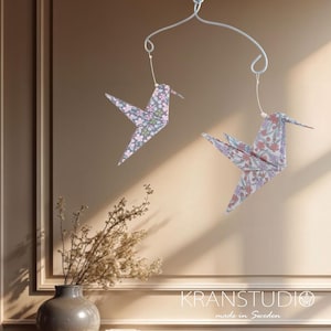 May include: Two origami hummingbird mobiles with floral patterns suspended from a silver wire frame. The birds feature pink, purple, and green flowers on a grey background. The mobile hangs in front of a neutral-toned wall.