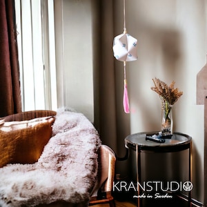 Peut inclure: Une lanterne en papier géométrique blanche avec un pompon rose suspendu au plafond dans une pièce avec un fauteuil marron et une table d'appoint avec un vase de fleurs séchées. La lanterne a un motif de feuilles bleues et blanches. Le texte "KRANSTUDIO made in Sweden" est visible dans le coin inférieur droit de l'image.