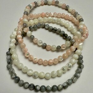 Può includere: Una collezione di cinque braccialetti di perline nei toni del grigio, rosa e bianco. I braccialetti sono fatti di piccole perline rotonde e sono disposti in un motivo circolare. Le perline hanno una lucentezza perlescente.