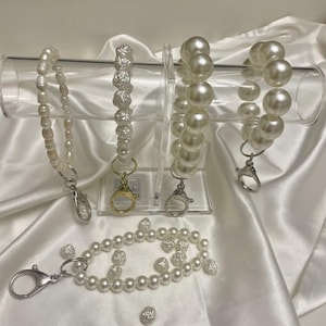 May include: Three pearl keychains with different styles: one with small pearls, one with larger pearls, and one with pearl roses. The keychains are displayed on a white acrylic stand. There are also loose pearl beads on a white surface.