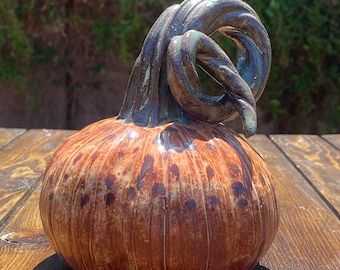 Può includere: Una zucca in ceramica con una glassa marrone e bianca. Il gambo è riccio e ha una glassa verde e marrone.