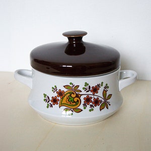 May include: A white ceramic serving dish with a brown lid and a floral design. The dish has two handles and is decorated with orange, green, and brown flowers.