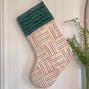 以下が含まれることがあります： 緑色のキルティングカフと、クリーム色の背景に赤、緑、金色の波線模様が施されたボディが特徴のクリスマスストッキング。白い壁に吊るされています。