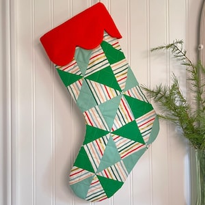 以下が含まれることがあります： 赤いスカラップトップとパッチワークデザインのクリスマスストッキング。パッチワークは、赤、黄、青など、さまざまな色の緑の三角形とストライプが特徴です。ストッキングは白い壁に掛けられています。