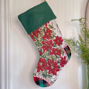 May include: Christmas stocking with a green cuff and a poinsettia floral pattern. The body of the stocking features red poinsettias and green leaves on a white background. The toe is made of red and black plaid fabric, trimmed with a candy cane stripe.