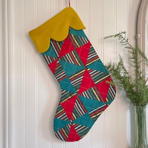 以下が含まれることがあります： 黄色のスカラップカフが付いたキルティングクリスマスストッキング。 赤とティールの三角形と金、白、黒のストライプの幾何学模様。 白いパネルの壁に掛けられています。