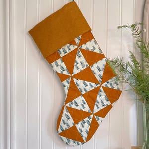 May include: A festive Christmas stocking with a patchwork design of orange and white triangles, featuring small green tree patterns. The cuff is a solid mustard colour. It hangs against a white panelled wall.
