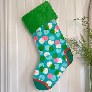 Peut inclure: Une chaussette de Noël matelassée avec un fond turquoise et des pois colorés verts, roses et blancs. Le revers et le bout sont verts, avec une bordure rouge. Accrochée à un mur blanc.