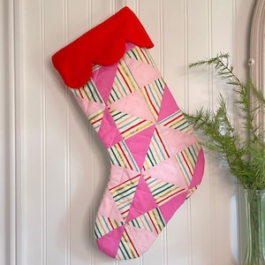 以下が含まれることがあります： 赤いスカラップカフが付いた、キルティング加工のクリスマスストッキング。ストッキングの本体はピンク色で、幾何学模様とカラフルなストライプが施されています。白い壁に掛けられています。