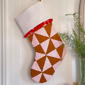 May include: A Christmas stocking with a white cuff and red pom-pom trim. The body features a geometric pattern of brown triangles and red-and-white striped triangles. A brown leather loop is attached for hanging.