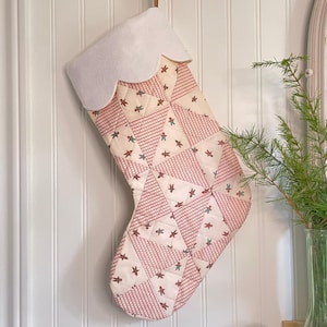 May include: A festive Christmas stocking with a white, scalloped top. The stocking is made from patchwork fabric in red and cream, decorated with star and gingerbread man patterns. It is suspended by a brown ribbon.