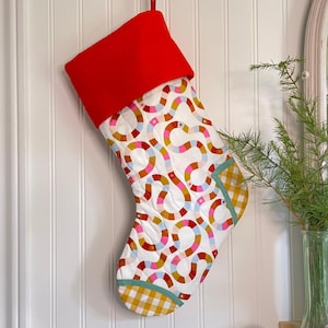 Puede incluir: Un calcetín navideño acolchado con puño rojo y cuerpo blanco estampado con formas coloridas entrelazadas. La puntera y el talón presentan un estampado de cuadros vichy amarillo y blanco, con un borde turquesa. Se adjunta un lazo rojo para colgar.