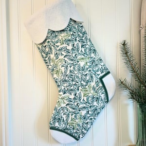 May include: A Christmas stocking with a white scalloped cuff and a green and white holly leaf pattern. The stocking has a white toe and heel with a green border. The stocking is hanging on a white paneled wall.