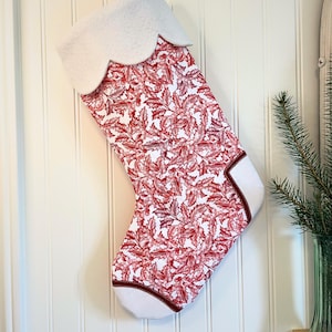 May include: A Christmas stocking with a white scalloped cuff and a red and white floral pattern. The stocking has a white toe and heel with a red border. The stocking is hanging on a white wall.