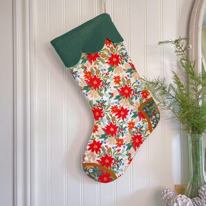 May include: A Christmas stocking with a green scalloped top and a floral pattern of red poinsettias, orange flowers, and green leaves on a white background. The stocking is hanging on a white wall.