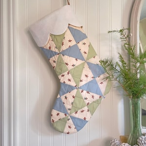 Puede incluir: Un calcetín navideño acolchado con un puño blanco y un diseño de patchwork. El patchwork presenta triángulos de tela azul claro, verde y crema, decorados con pequeñas figuras rojas de hombre de jengibre. El calcetín cuelga de una pared blanca.