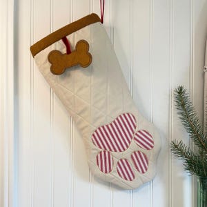 May include: Cream-coloured Christmas stocking with a quilted pattern and a brown felt dog bone appliqué. The paw-shaped design features red and white striped fabric. A red ribbon loop is attached for hanging.