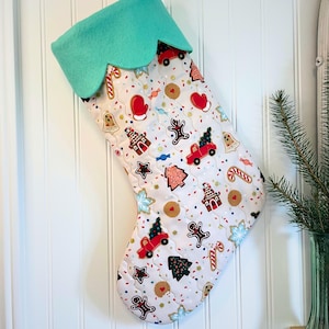 Peut inclure: Une chaussette de Noël avec un haut festonné bleu sarcelle clair. Le corps de la chaussette est blanc avec un motif matelassé et présente un motif répété de bonhommes en pain d'épices, d'arbres et de cannes en sucre. Des camions et des mitaines rouges sont également inclus.