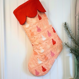 May include: A quilted Christmas stocking with a peach background and a pattern of white, pink, and red fir trees. The top of the stocking is red felt with a scalloped edge. Gold star accents are scattered across the stocking.