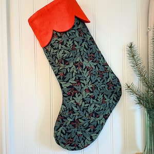 May include: A Christmas stocking with a black background featuring a pattern of green leaves and red berries. The top is bright orange with a scalloped edge. It hangs on a white wall. The stocking is approximately 50 cm long.