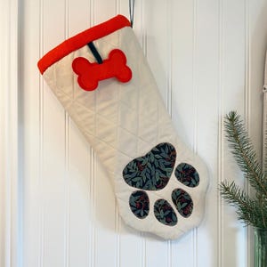 May include: Cream quilted Christmas stocking with a red bone applique and a paw print design. The paw print is made of dark blue fabric with a green leaf and red berry pattern. The top of the stocking is trimmed with red fabric.