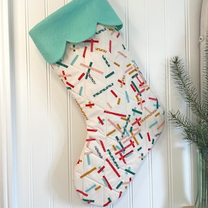 May include: A quilted Christmas stocking featuring a white base with a scattered pattern of small, multi-coloured rectangular shapes in red, teal, yellow, and pink. The stocking has a solid mint green cuff with a scalloped bottom edge.