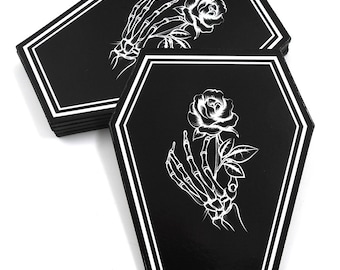 Può includere: Set di sottobicchieri neri a forma di bara con contorni bianchi. Ogni sottobicchiere presenta una mano scheletrica bianca che tiene una rosa bianca.