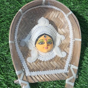 May include: A decorative, handcrafted piece featuring a painted face with a serene expression, set within a woven bamboo frame. The face is adorned with a white headdress and delicate accents. The frame is supported by small legs.