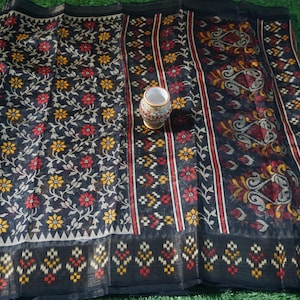 May include: A black textile with floral and geometric patterns, featuring red and yellow flowers. A small, decorative vase with a patterned design sits on the fabric. The textile has a dark border with a repeating geometric design.