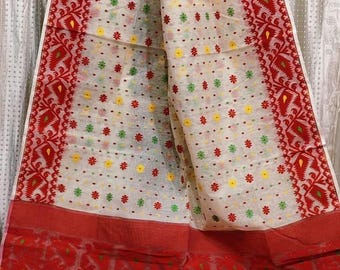 May include: A cream-colored saree featuring a wide red border with intricate geometric patterns. The main body is decorated with small, repeating floral motifs in red, yellow, and green. A solid red panel is at the bottom with yellow accents.