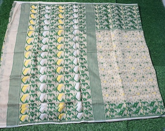 May include: A beige textile with green and yellow floral patterns. The fabric has a green border and is laid out on a green surface. The design includes repeating floral and geometric shapes.