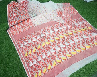 May include: A patterned textile with a repeating design of red, yellow, and white shapes on a white background. The textile has a red border and is folded on a green surface.