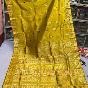 May include: A mustard yellow saree featuring a wide gold border with intricate woven patterns of figures and floral motifs. The main body of the fabric displays small, scattered red and gold floral designs throughout the length of the garment.