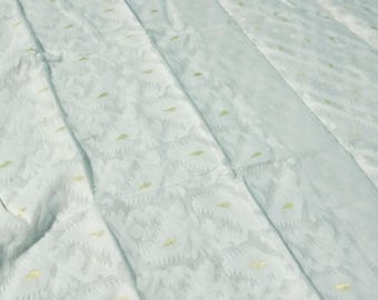 May include: A light blue fabric with a repeating floral pattern and small yellow-green accents. The fabric is folded, creating visible lines and shadows across the surface. The overall texture appears soft and slightly translucent.