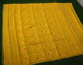 May include: A bright yellow textile with a repeating pattern of small red diamond shapes. The fabric is folded and laid out on a green surface, showcasing its texture and design. The textile has a detailed border.