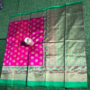 May include: A bright pink saree with gold floral patterns and a green border. The saree has a decorative green and gold patterned section. A small, ornate white and gold container rests on the fabric.