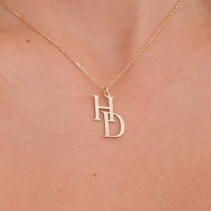 Puede incluir: Un delicado collar dorado con un colgante entrelazado "HD". El colgante cuelga de una fina cadena, creando un aspecto minimalista y elegante. El collar se muestra sobre un fondo de tono de piel.