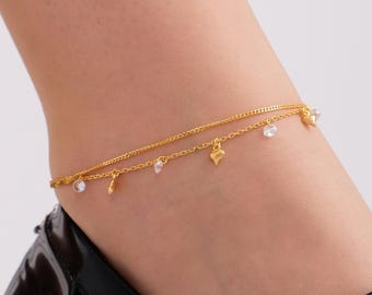 May include: A gold-colored double-layered anklet with small heart and clear crystal charms. The anklet is worn on the ankle. The jewelry is delicate and elegant.