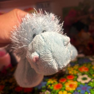 May include: A light blue plush toy featuring a shaggy, textured mane, small dark eyes, and tiny pink accents on the snout. The stuffed animal has a soft, fuzzy exterior and a rounded, prominent muzzle, crafted from synthetic fabric materials.
