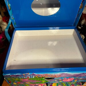 Puede incluir: Una caja de madera azul con tapa con bisagras y una abertura circular. El interior es blanco y la caja est&aacute; decorada con una colorida escena de paisaje con un castillo y el nombre "Cassara". La caja tiene un asa a cada lado.