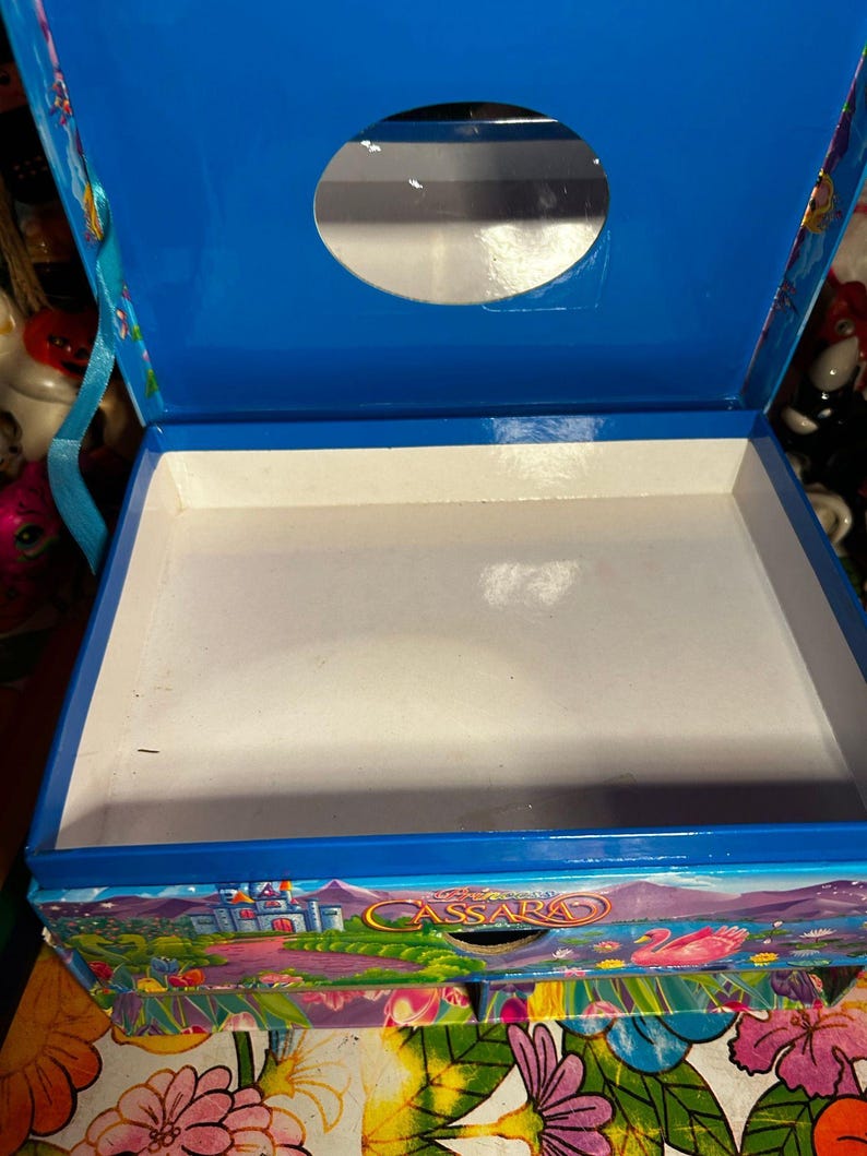Puede incluir: Una caja de madera azul con tapa con bisagras y una abertura circular. El interior es blanco y la caja est&aacute; decorada con una colorida escena de paisaje con un castillo y el nombre "Cassara". La caja tiene un asa a cada lado.
