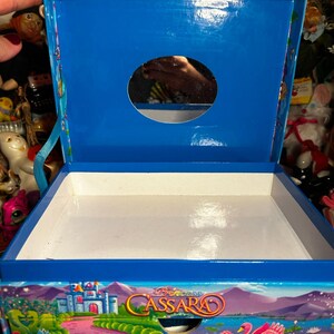 Puede incluir: Caja azul abierta con interior blanco. La caja presenta una colorida ilustraci&oacute;n de un castillo, un cisne rosa y el texto "Princess Cassara". La parte superior de la caja tiene una abertura circular.