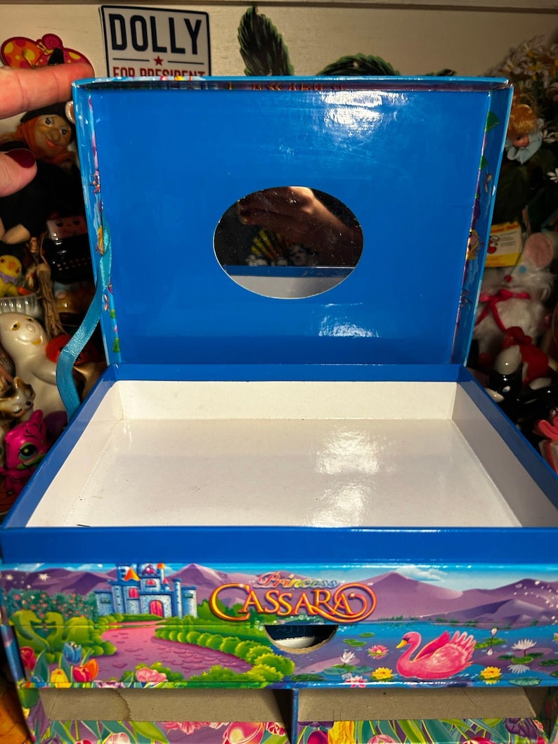 Puede incluir: Caja azul abierta con interior blanco. La caja presenta una colorida ilustraci&oacute;n de un castillo, un cisne rosa y el texto "Princess Cassara". La parte superior de la caja tiene una abertura circular.