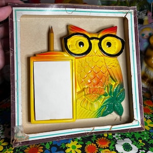 May include: A vintage owl-shaped memo holder with a pencil and a blank notepad. The owl is yellow with black glasses and orange accents. The holder is in a brown and white box, with the text "DOLLY FOR PRESIDENT" visible in the background.