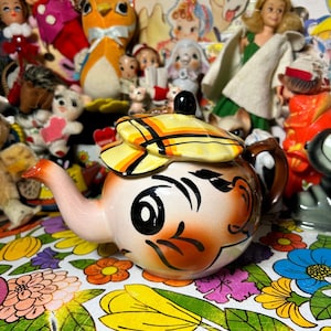 May include: A ceramic teapot shaped like a cartoon animal with a yellow and orange striped hat. The teapot is painted with brown stripes and has a black eye and a black nose. The teapot is sitting on a floral tablecloth.