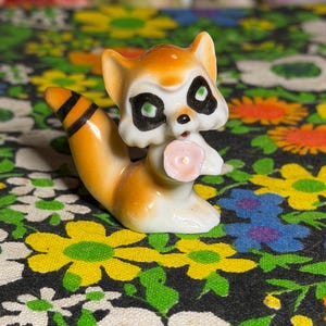 Puede incluir: Una pequeña figura de mapache de cerámica con pelaje naranja y blanco, parches negros alrededor de los ojos y una cola rayada. El mapache sostiene una diminuta flor de color rosa pálido en sus patas delanteras, con un acabado brillante.